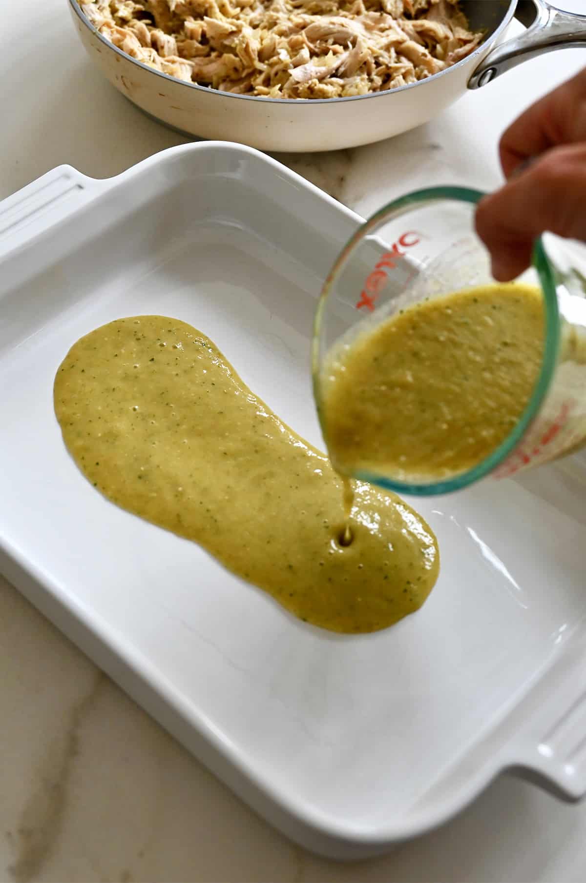 Pouring green salsa from a liquid measuring cup into a baking dish.