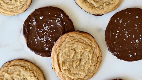 Chocolate chipless cookies, some dipped in chocolate and sprinkled with sea salt, on a marble countertop.