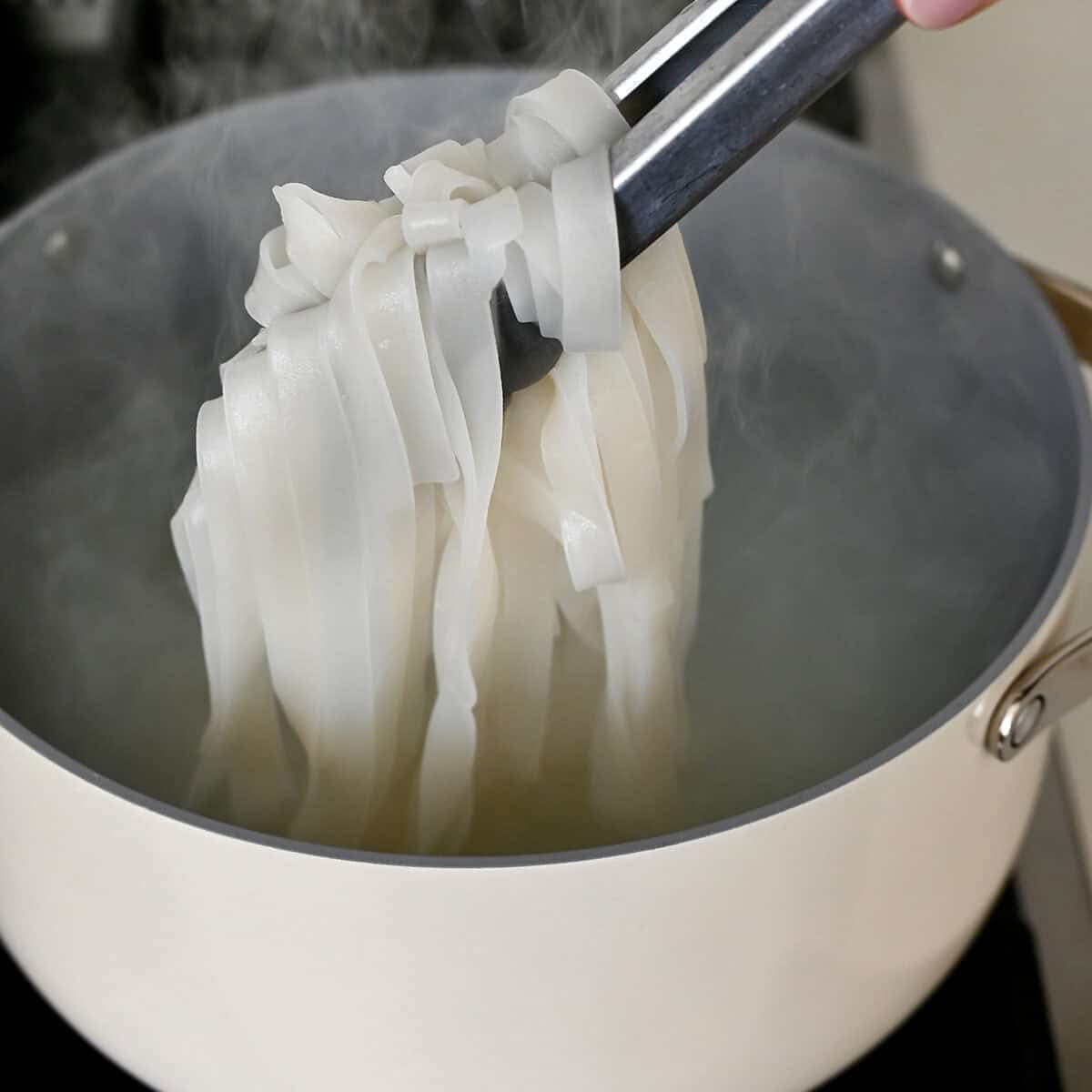 Cooked wide rice noodles being held out of a pot of water with tongs.