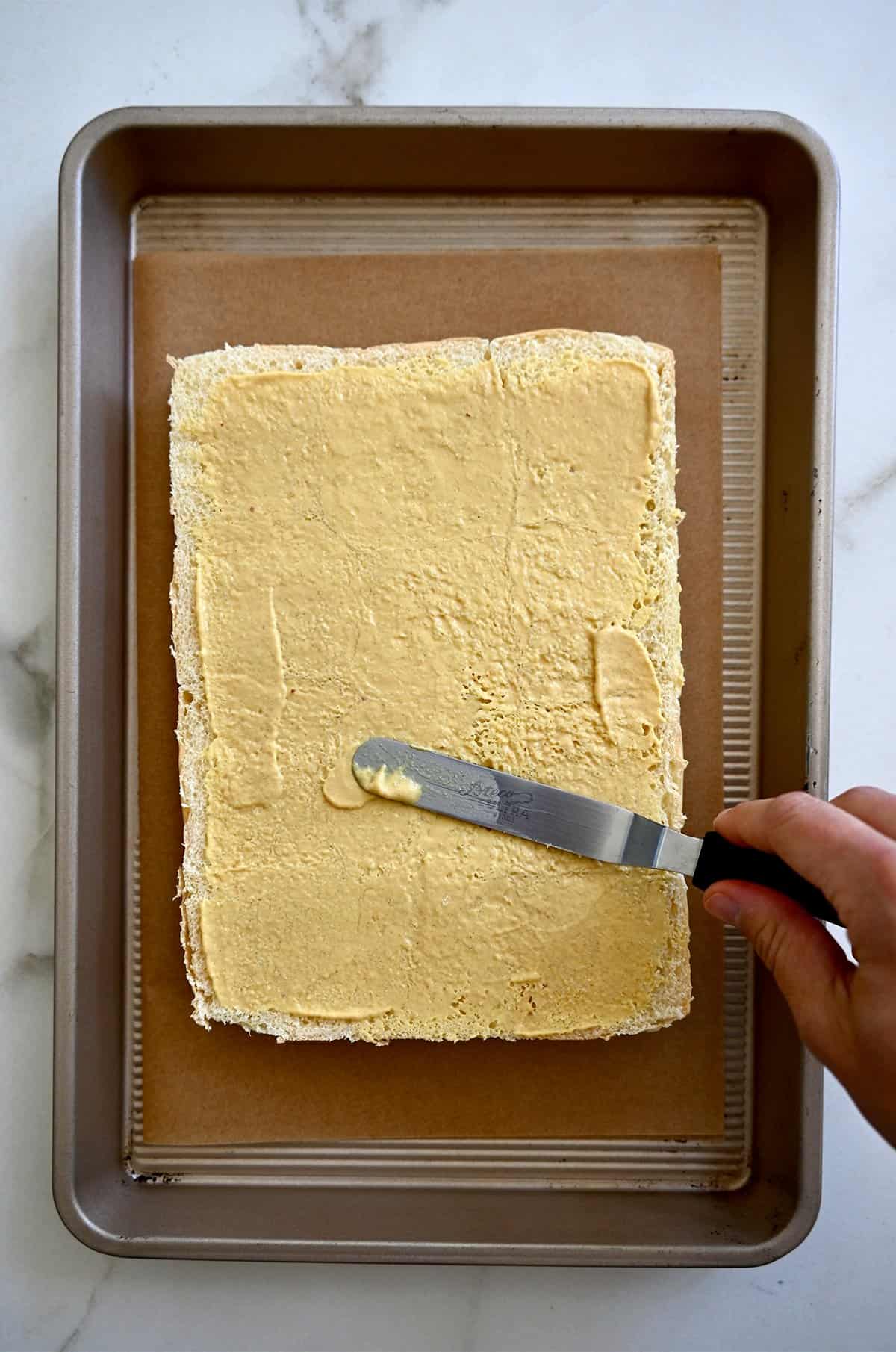 A hand holding an offset spatula smears Dijon mustard atop the bottom half of slider rolls.
