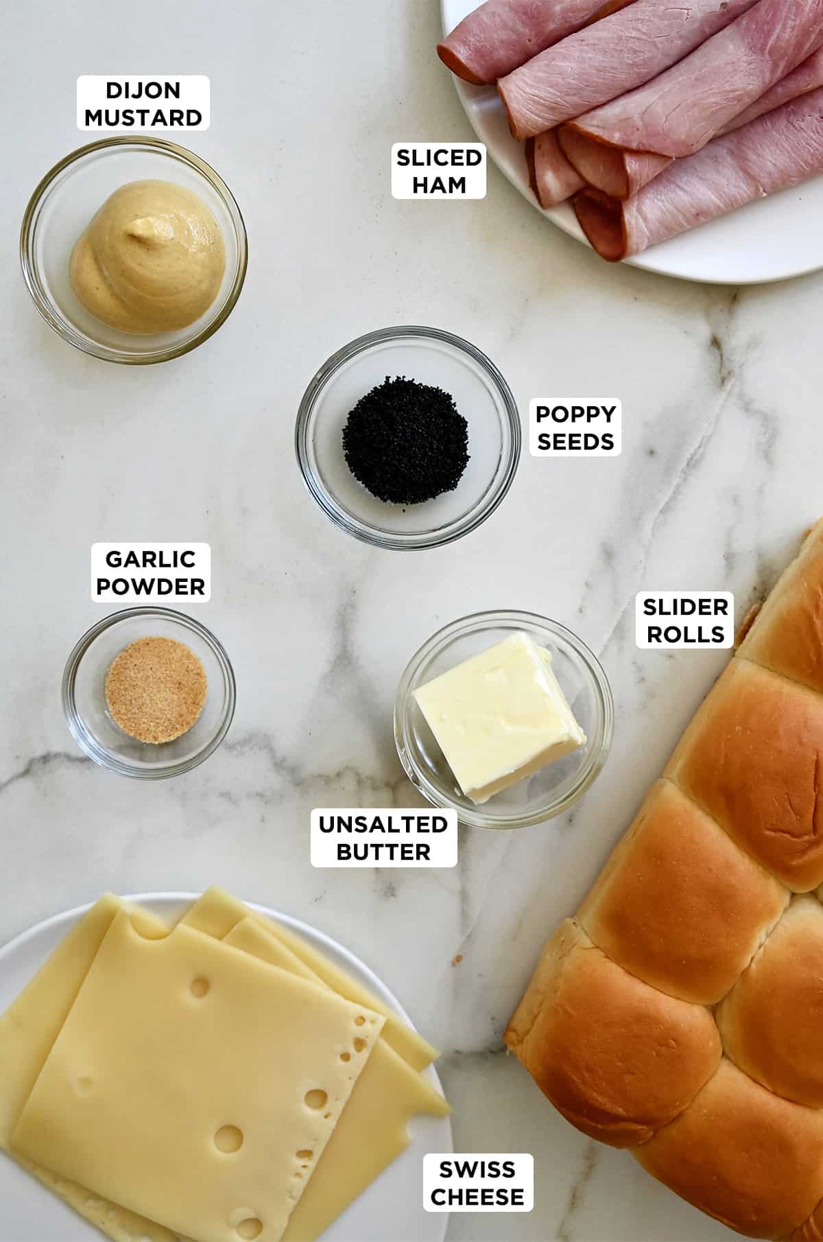 Sweet slider rolls next to a plate with Swiss cheese, a plate with deli ham, and bowls containing Dijon mustard, butter, garlic powder and poppy seeds.