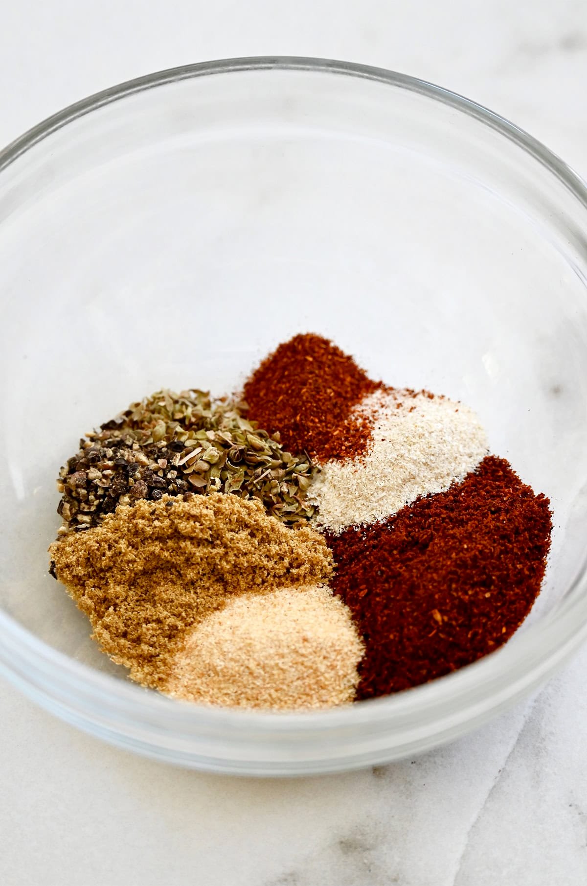 A glass bowl containing garlic powder, garlic onion, chili powder, ground cumin, black pepper, dried oregano and paprika