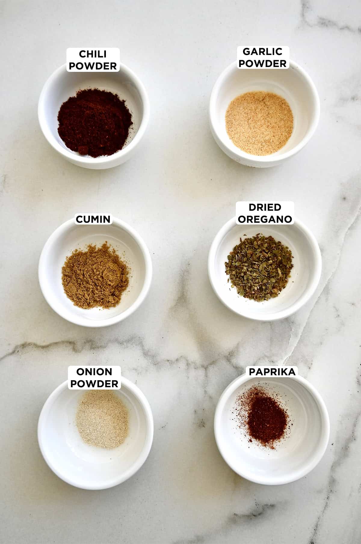 Six small bowls containing taco seasoning ingredients, including chili powder, ground cumin, onion powder, paprika, dried oregano and garlic powder.