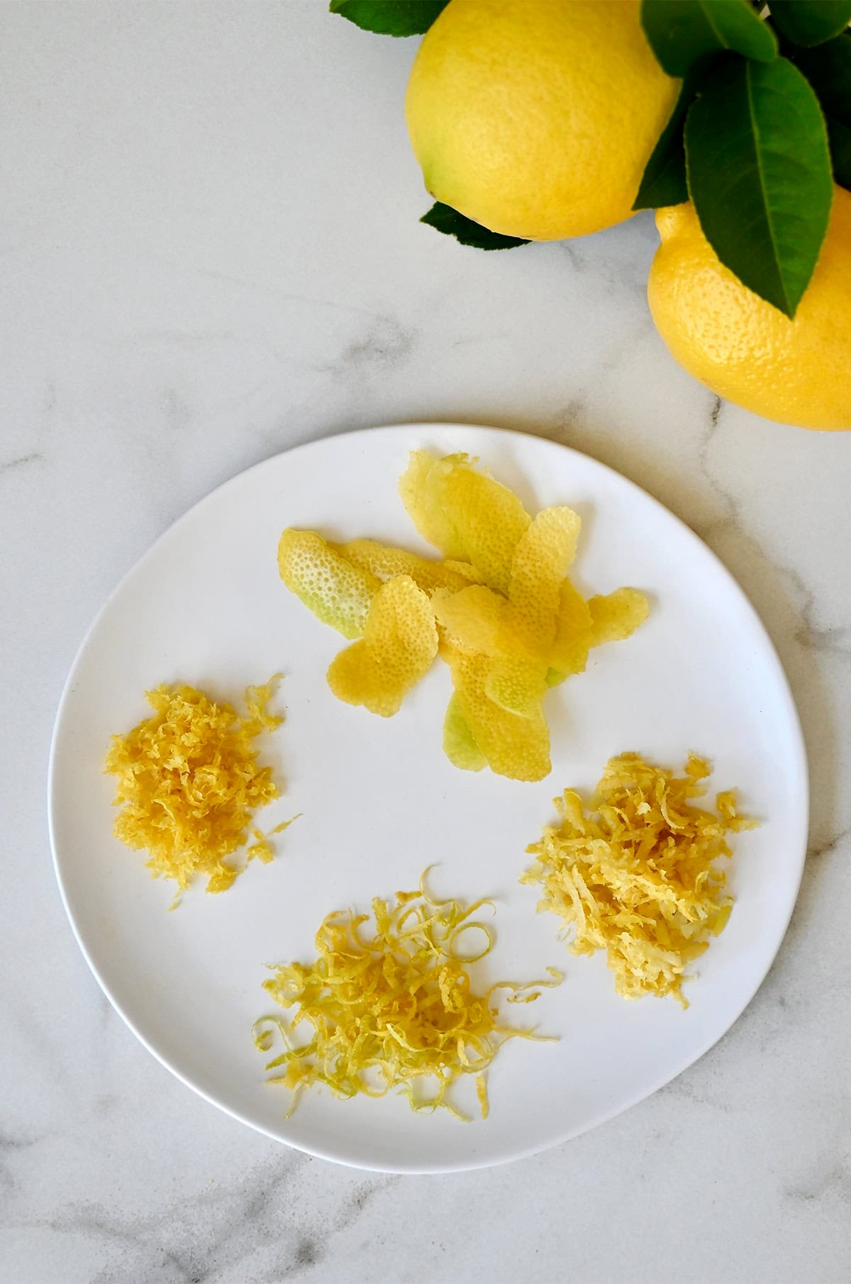 A plate with lemon zest that shows zest from four different techniques: A citrus zester, a microplane, a box grater and a vegetable peeler.