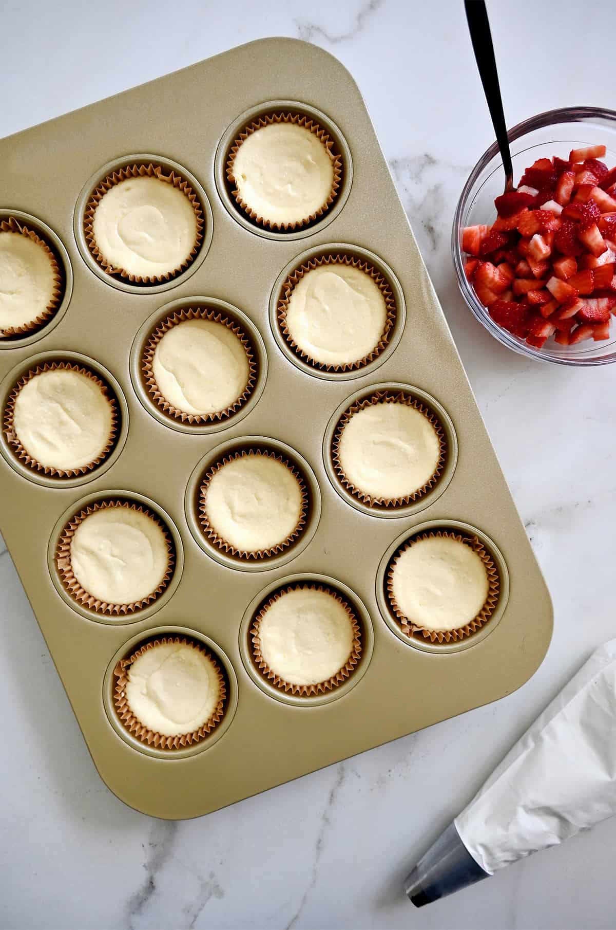 Baked mini cheesecakes in a muffin pan next to a bowl containing diced fresh strawberries.
