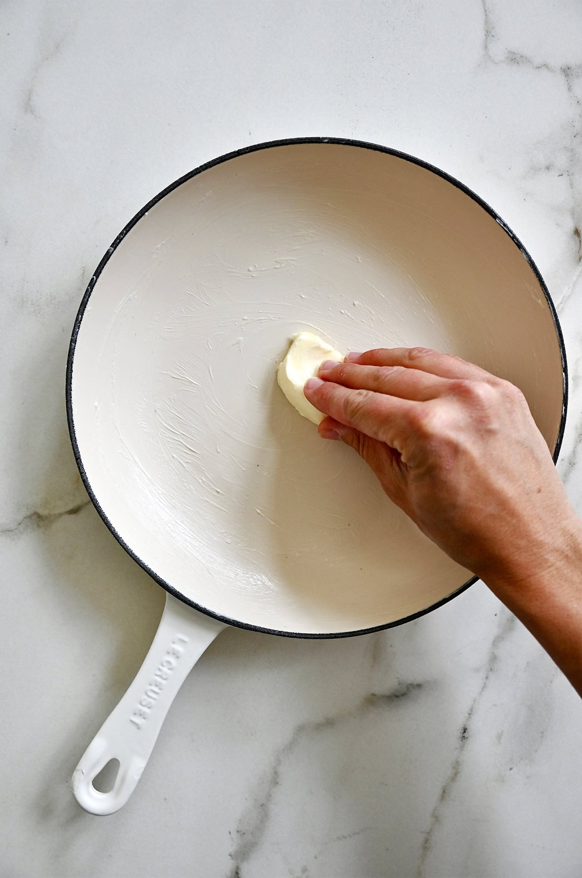 A hand greases an enameled cast iron skillet with butter.