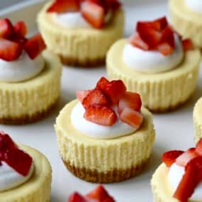 Mini cottage cheese cheesecakes topped with a dollop of whipped cream and chopped fresh strawberries on a serving plate.