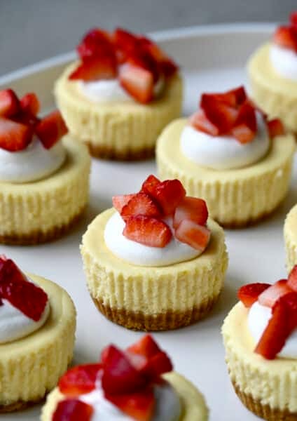 Mini cottage cheese cheesecakes topped with a dollop of whipped cream and chopped fresh strawberries on a serving plate.