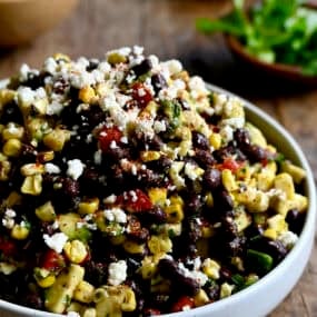A white serving bowl containing black bean and corn salad with avocado and cojita cheese all tossed in a tangy lime vinaigrette.