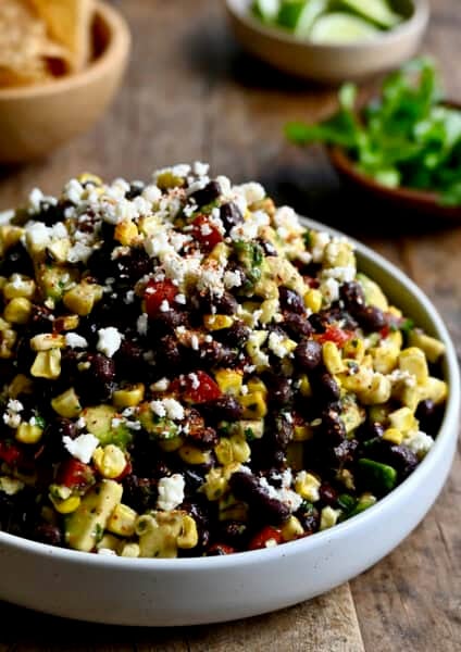 A white serving bowl containing black bean and corn salad with avocado and cojita cheese all tossed in a tangy lime vinaigrette.