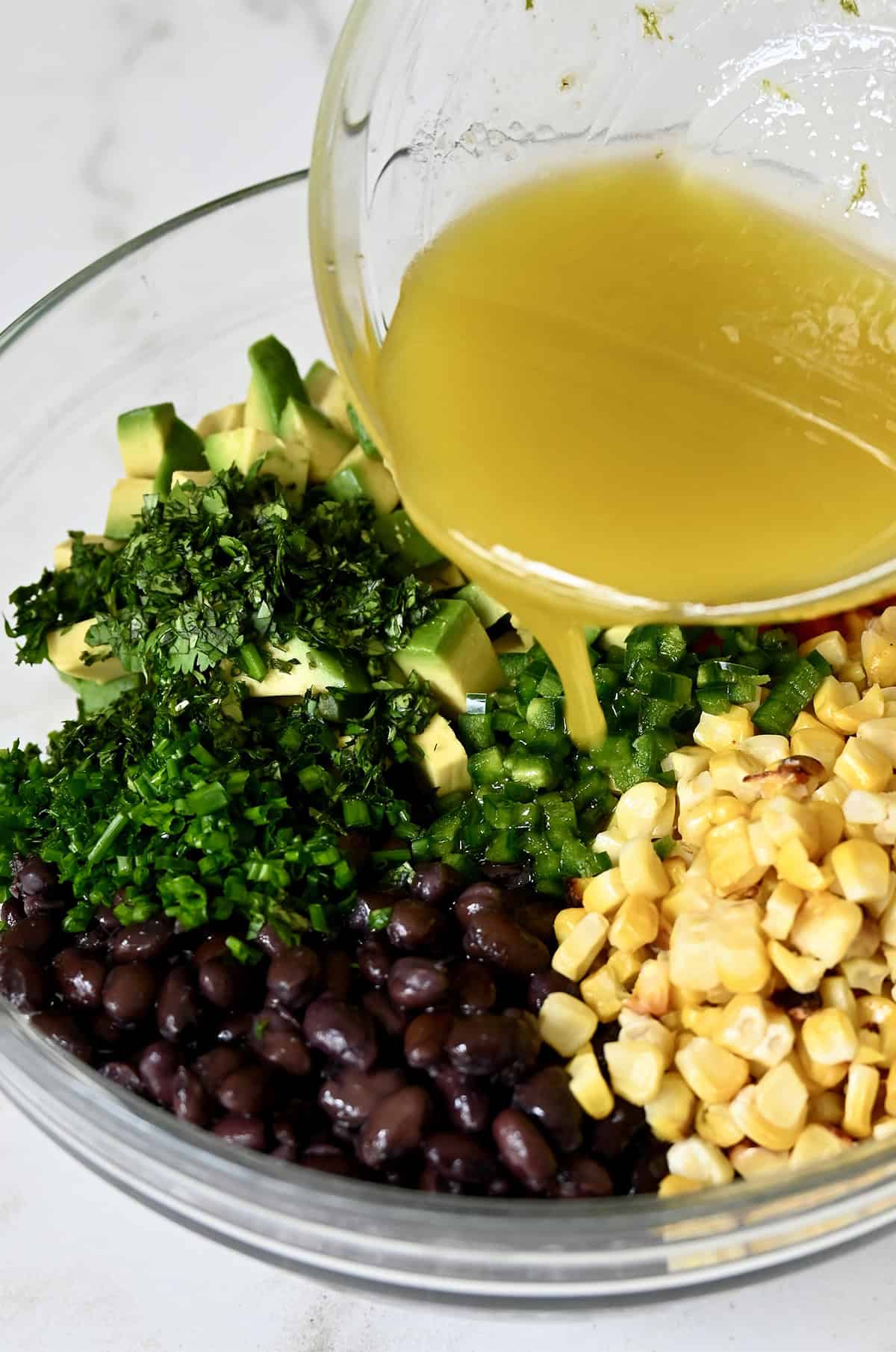 Pouring the lime vinaigrette from a small bowl over the corn, black bean and avocado mixture in another bowl.