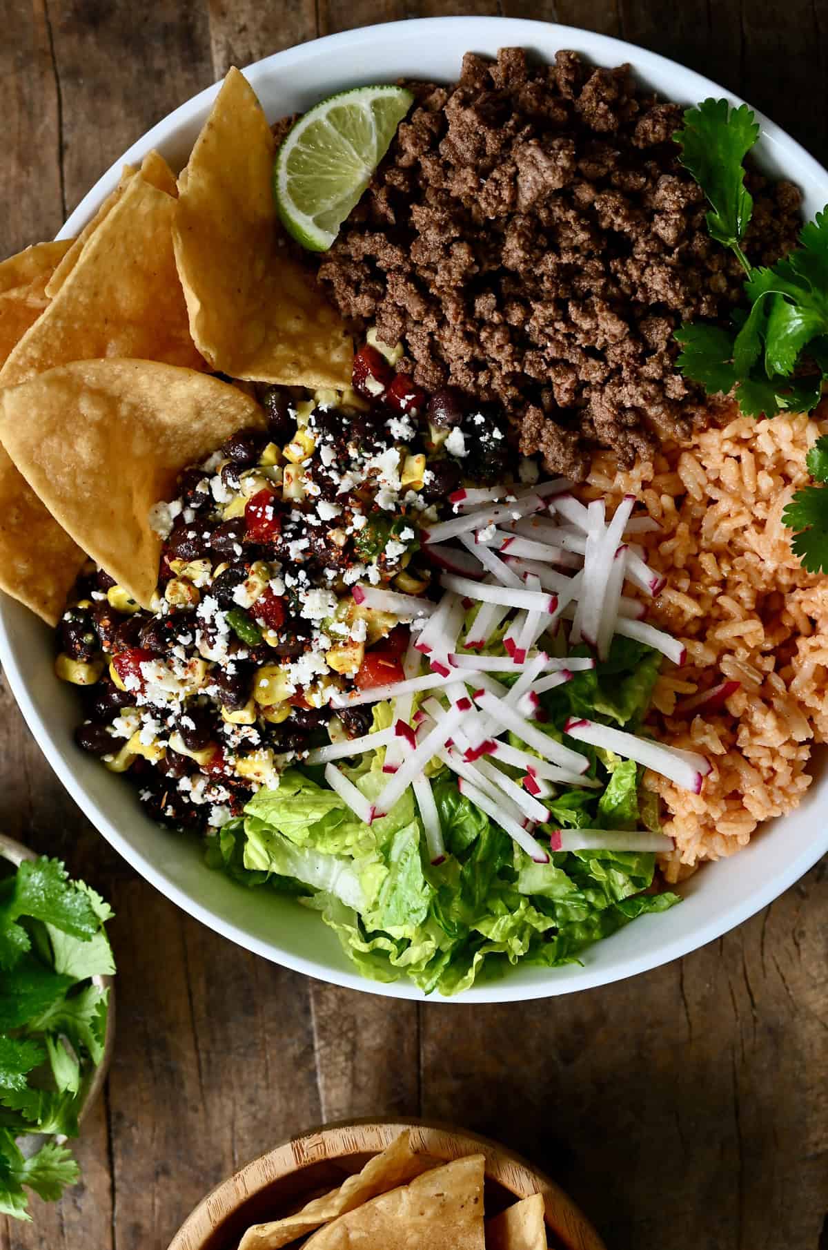 A taco bowl with shredded lettuce, ground beef, sliced radishes, black bean and corn salad, cilantro, and tortilla chips.