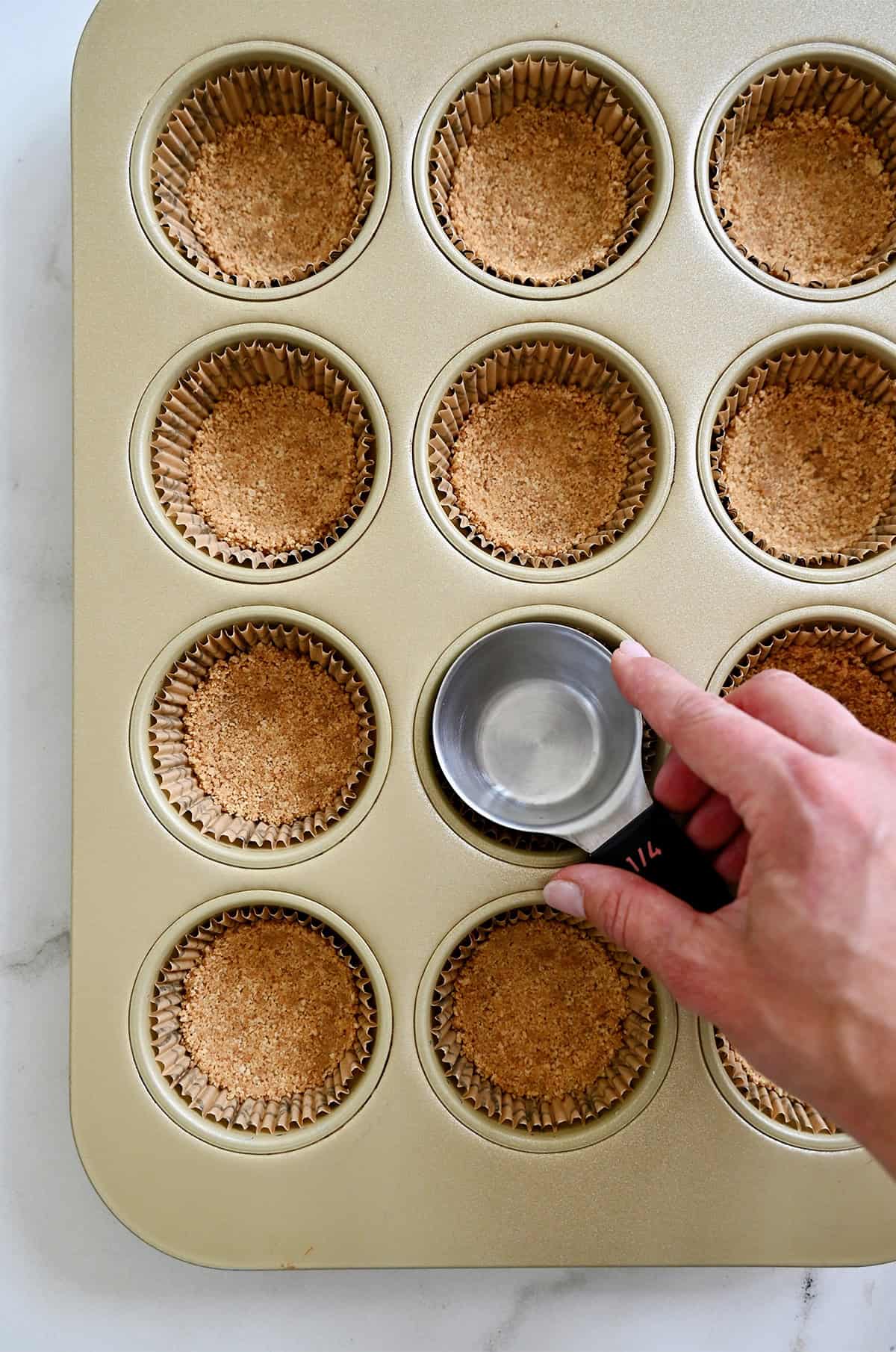 A hand holding a measuring cup presses crushed graham crackers into cups of a muffin tin to form cheesecake crust.