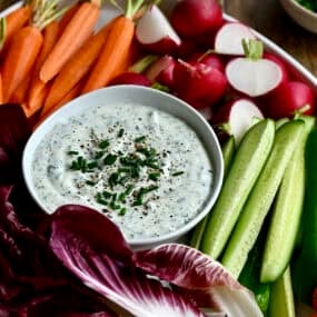 Cottage cheese dip in a bowl on a platter surrounded by fresh carrots, radishes, cucumbers and cherry tomatoes.