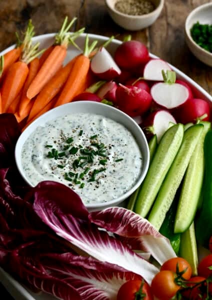 Cottage cheese dip in a bowl on a platter surrounded by fresh carrots, radishes, cucumbers and cherry tomatoes.