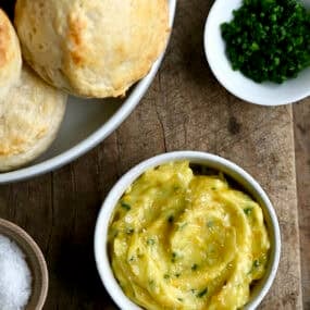 Homemade honey butter in a small bowl next to a bowl piled high with flaky buttermilk biscuits.