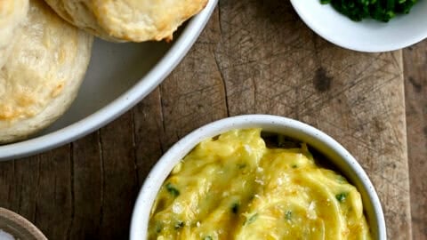 Homemade honey butter in a small bowl next to a bowl piled high with flaky buttermilk biscuits.