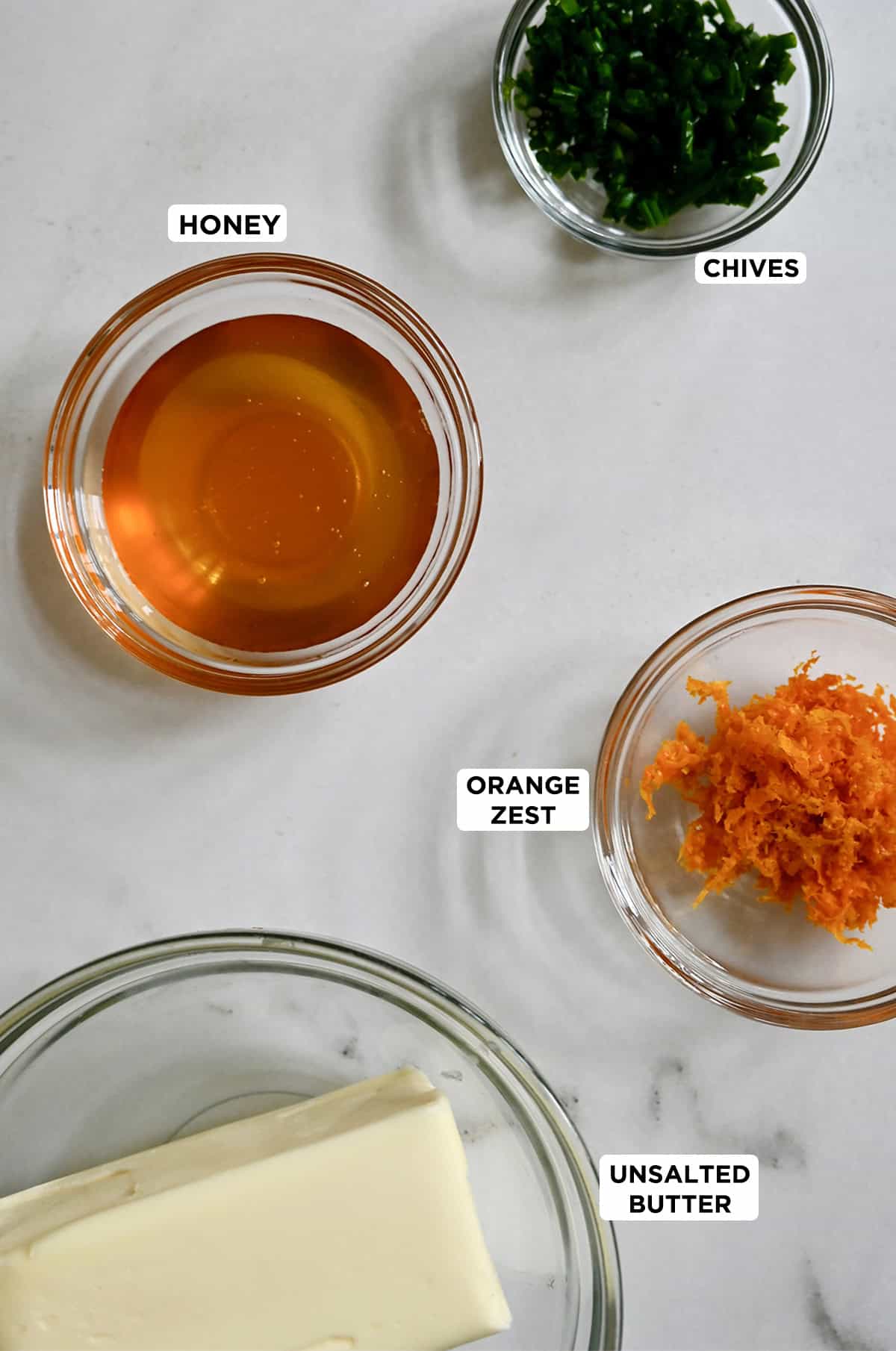 A bowl containing room temperature butter next to other bowls containing honey, chopped fresh chives and orange zest.