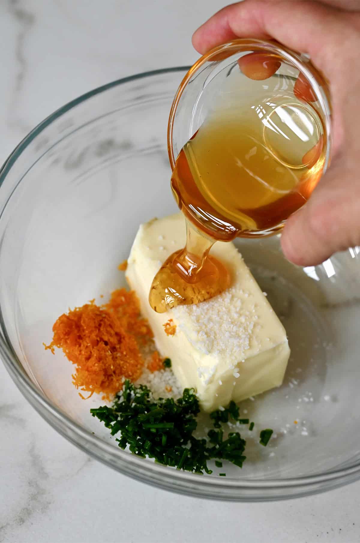 Honey being poured from a small bowl into a bigger bowl with softened butter, chopped fresh chives, salt and orange zest.