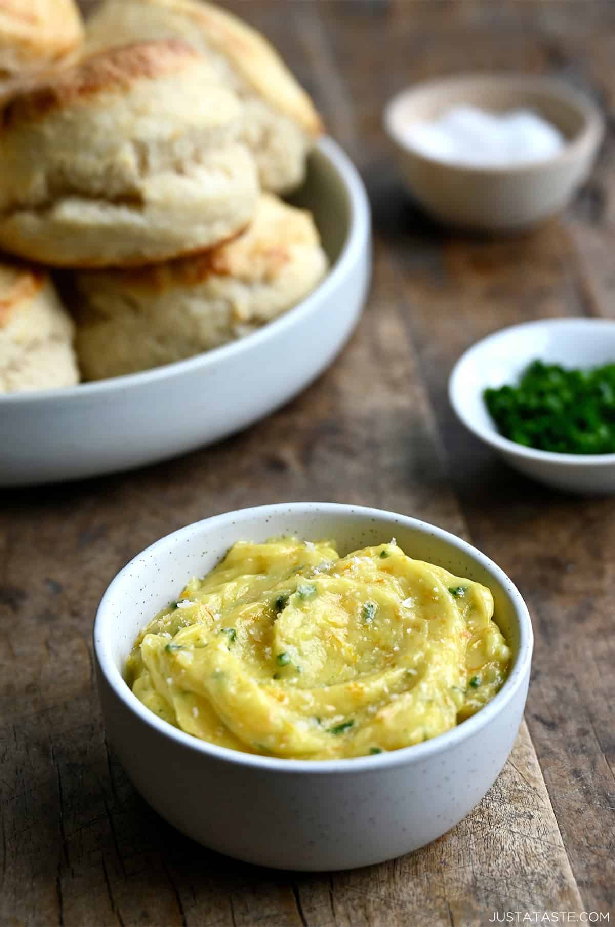 A small bowl containing honey butter with chopped fresh chives and orange zest near a bowl containing biscuits.