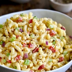 A serving bowl containing macaroni salad with diced red onion, diced celery and diced red bell pepper, all tossed in a creamy homemade dressing.