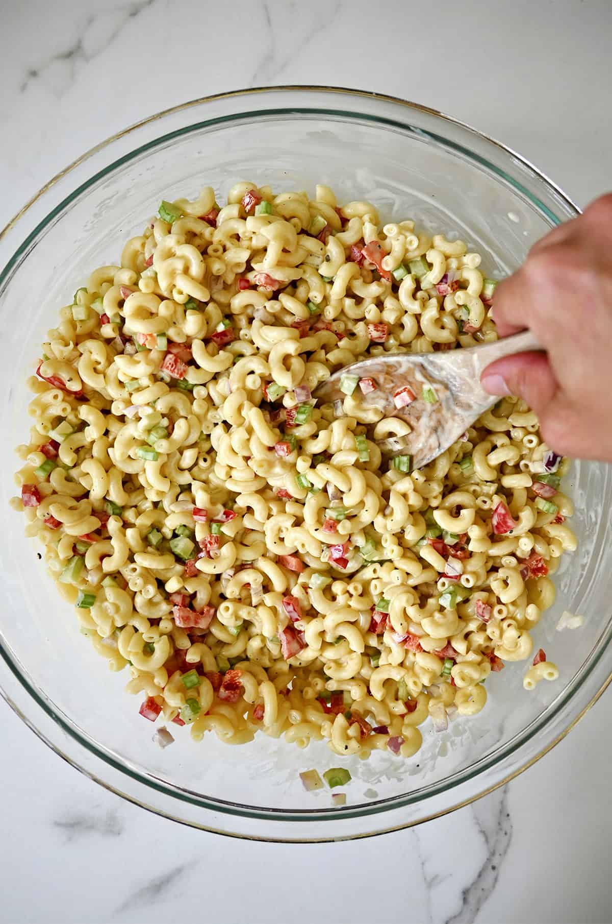 A hand holding a wooden spoon mixes macaroni salad with a creamy dressing in a large bowl.
