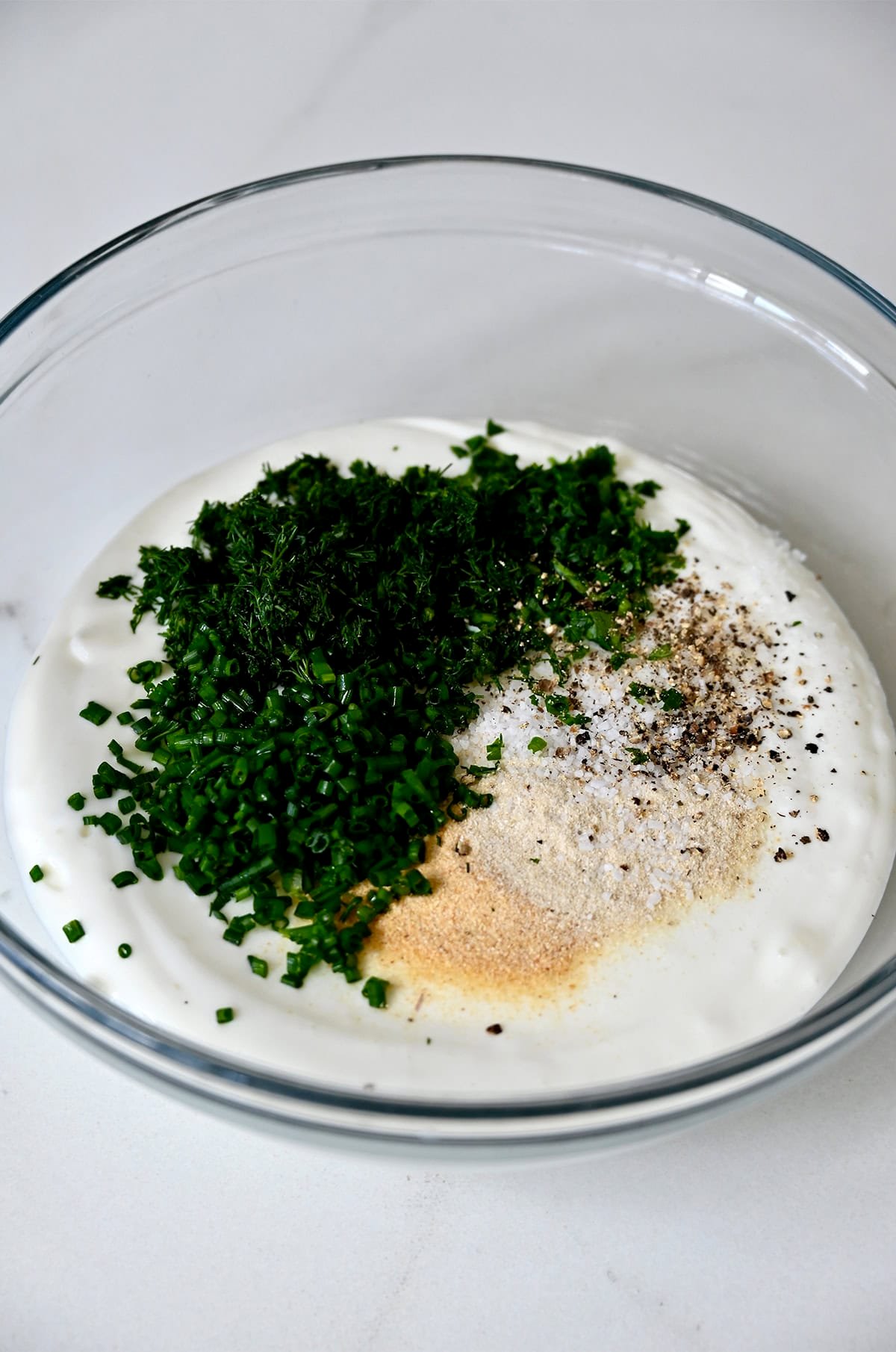 A glass bowl containing chopped fresh chives, parsley and dill, along with onion powder and garlic powder atop whipped cottage cheese. 