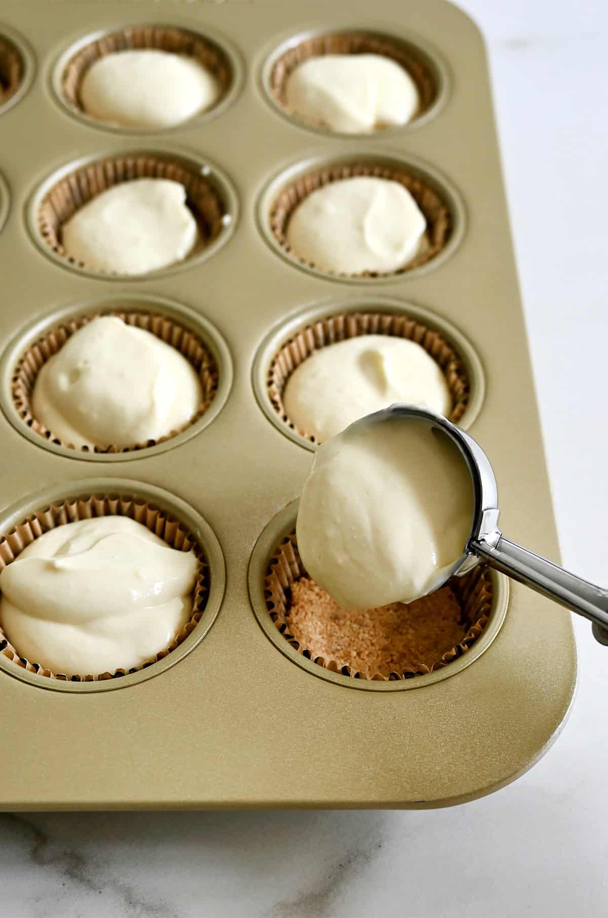 Spooning cottage cheese cheesecake batter atop graham cracker crust in a muffin pan.