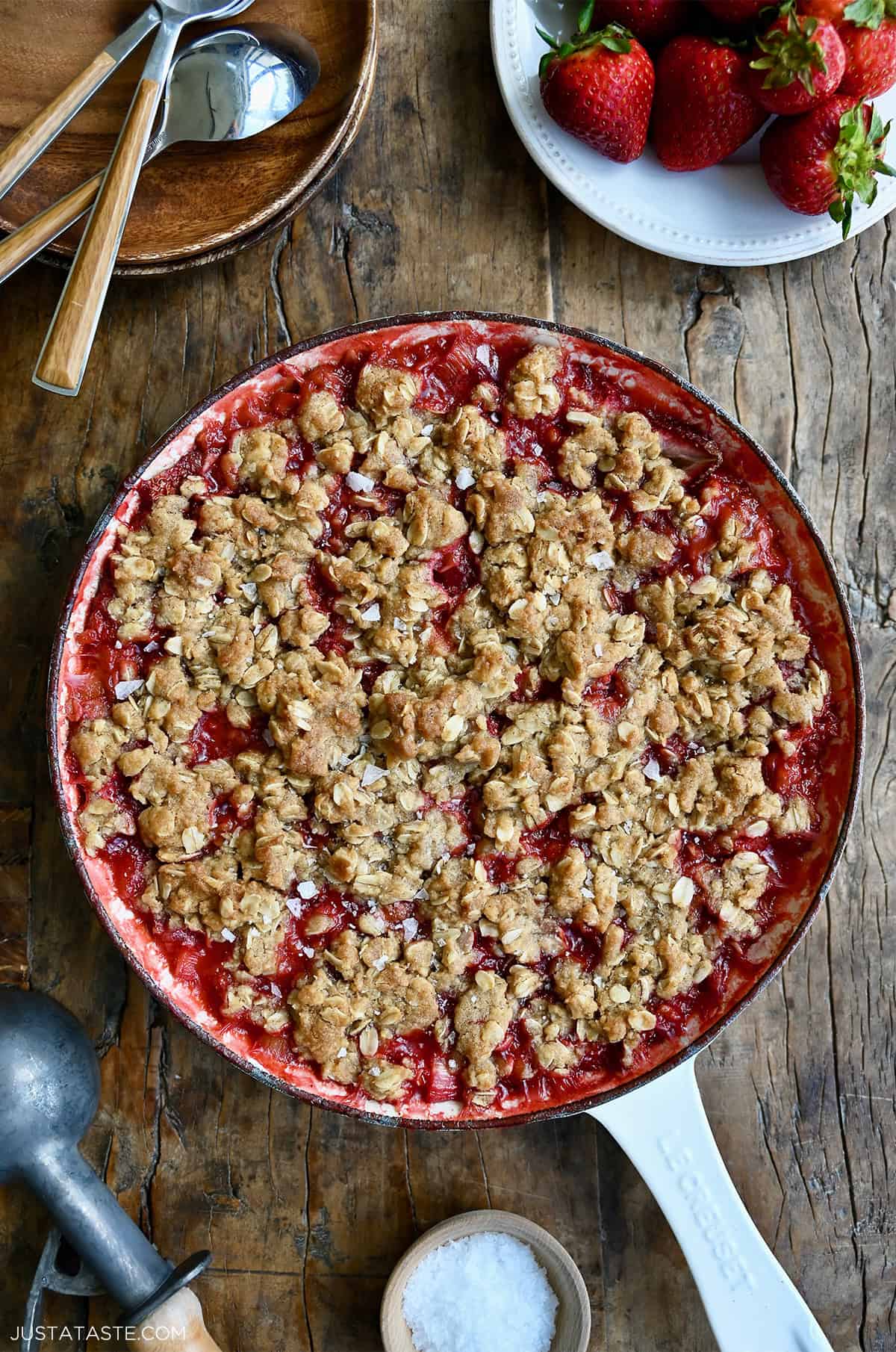 A white enameled cast iron skillet containing strawberry rhubarb crisp. A bowl with fresh strawberries and an ice scoop are nearby.