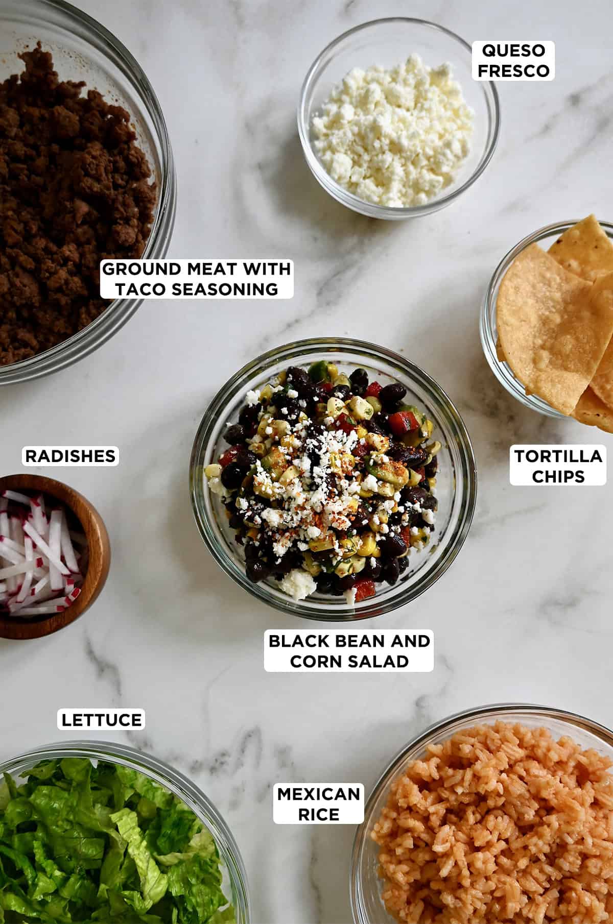 Bowls containing ground beef with taco seasoning, queso fresco, tortilla chips, black bean and corn salad, Mexican rice, shredded lettuce, and sliced radishes. 