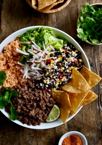 Taco meat in a bowl with Mexican rice, shredded lettuce, black bean and corn salad, sliced radishes, cilantro, a wedge of lime, and tortilla chips.