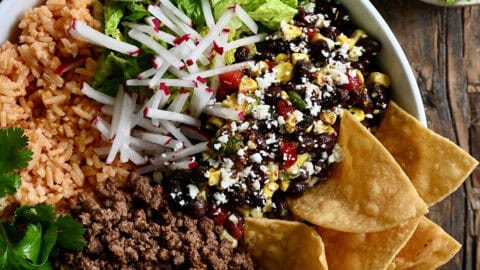 Taco meat in a bowl with Mexican rice, shredded lettuce, black bean and corn salad, sliced radishes, cilantro, a wedge of lime, and tortilla chips.