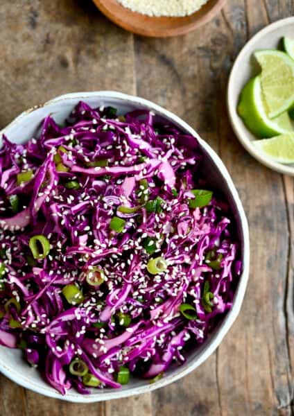 Asian slaw with sesame dressing in a salad bowl garnished with sesame seeds near a small bowl with lime wedges.