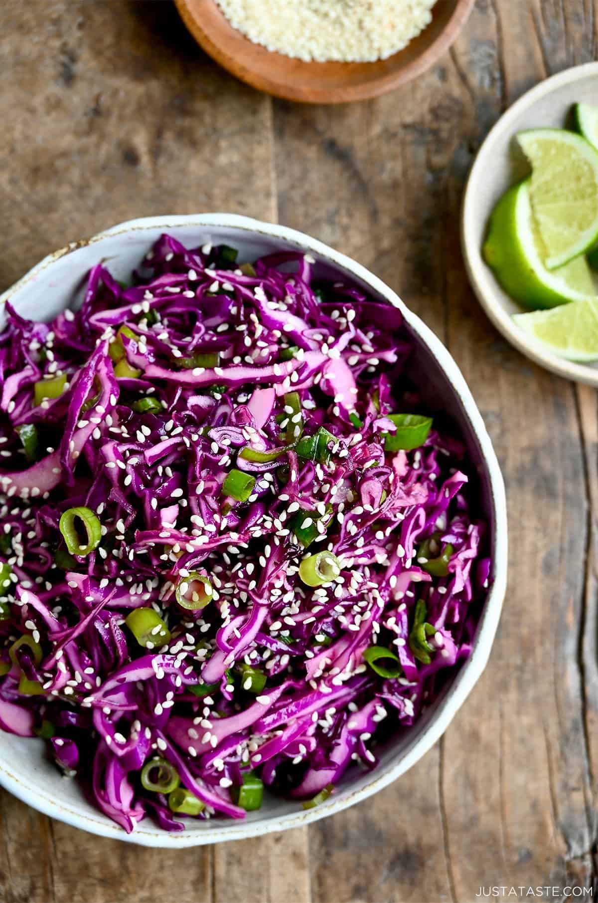 Asian slaw with sesame dressing in a salad bowl garnished with sesame seeds near a small bowl with lime wedges.