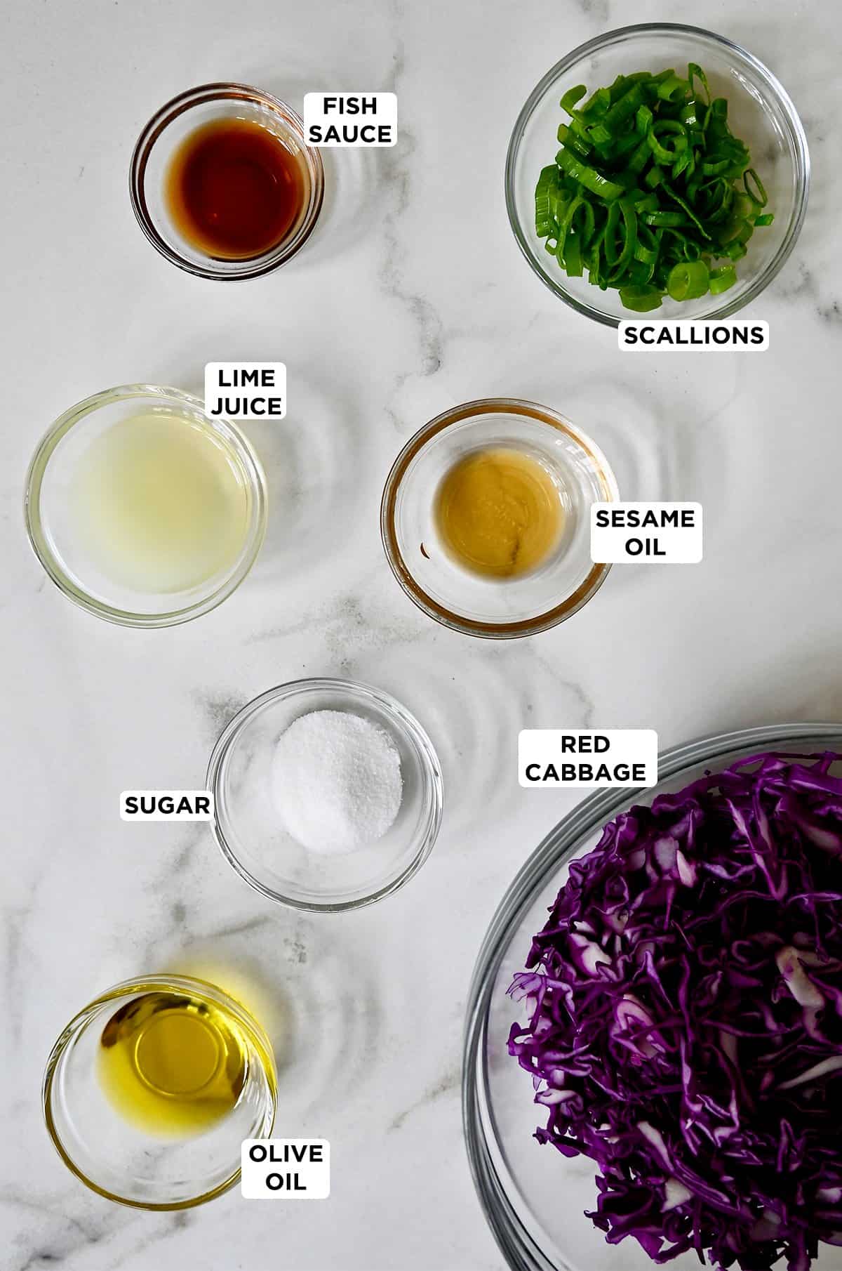 Asian slaw ingredients in various sizes of glass bowls, including shredded purple cabbage, olive oil, sugar, sesame oil, fish sauce, lime juice and chopped scallions.