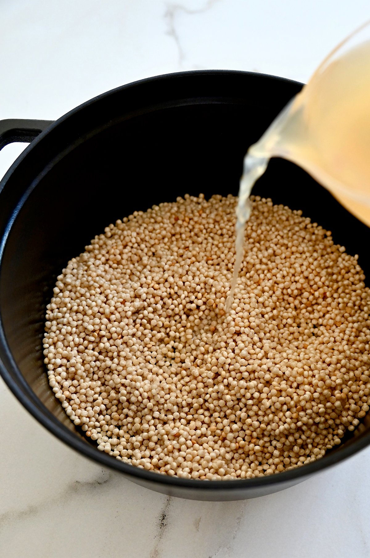 Adding chicken broth to a pot with toasted couscous.