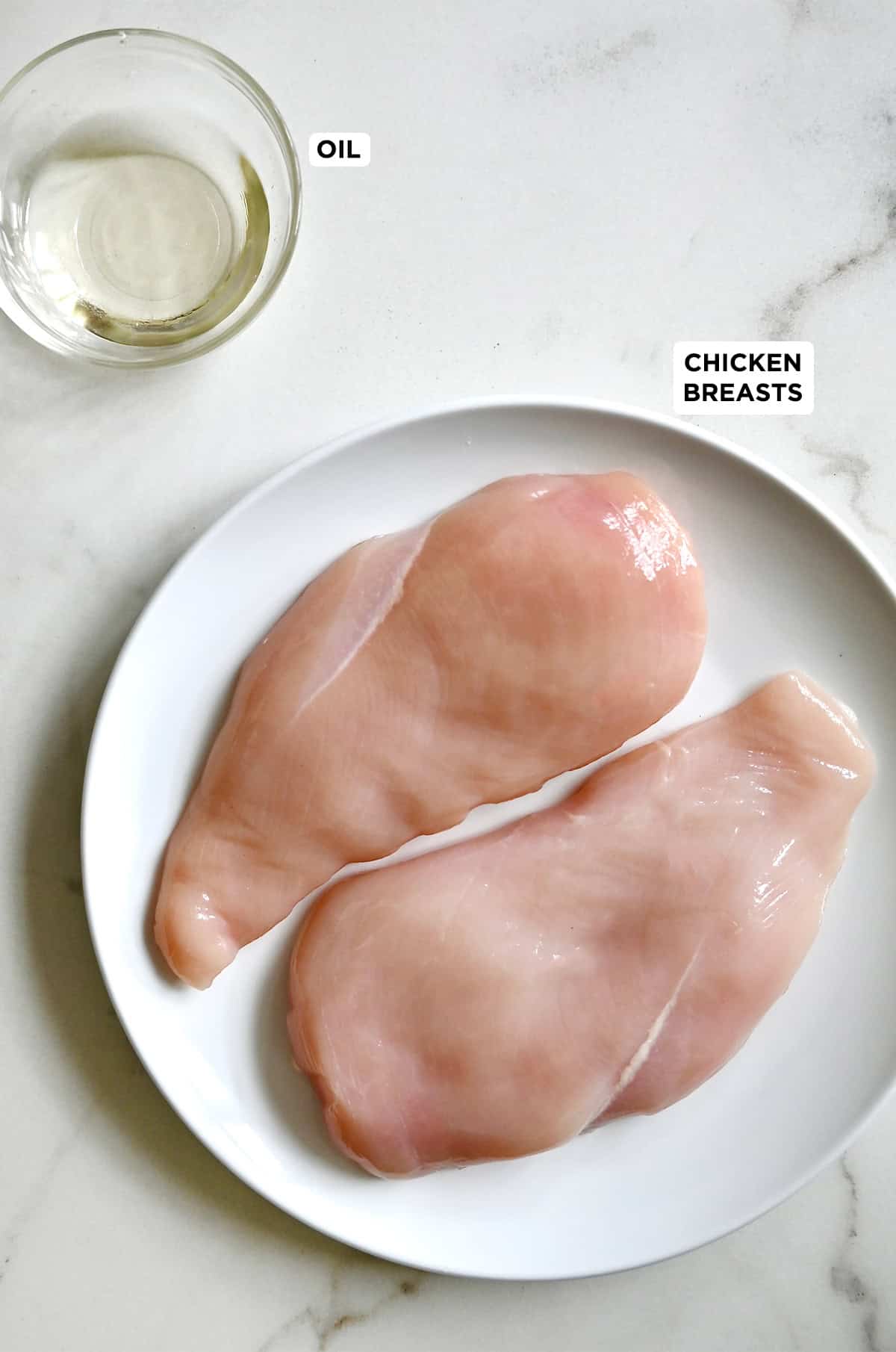 Two raw chicken breasts on a plate next to a small bowl containing oil.