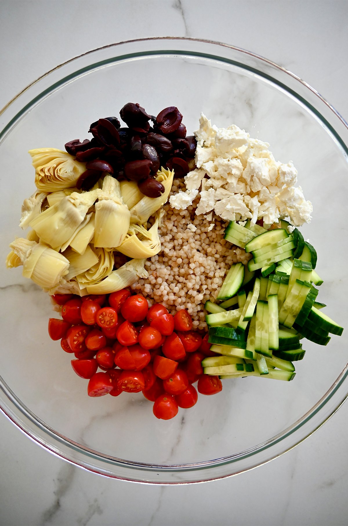 A large bowl containing cooked couscous, halved cherry tomatoes, artichoke hearts, sliced kalamata olives, crumbled feta cheese and sliced cucumbers.