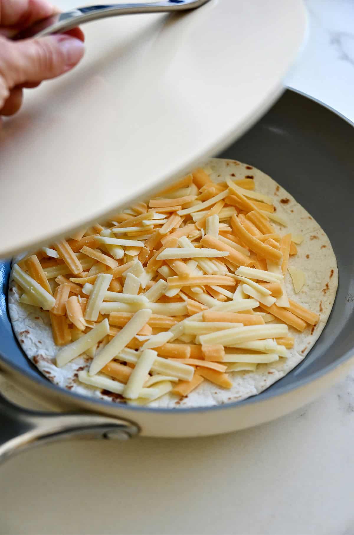 A tortilla topped with shredded cheese in a skillet being covered with a lid.