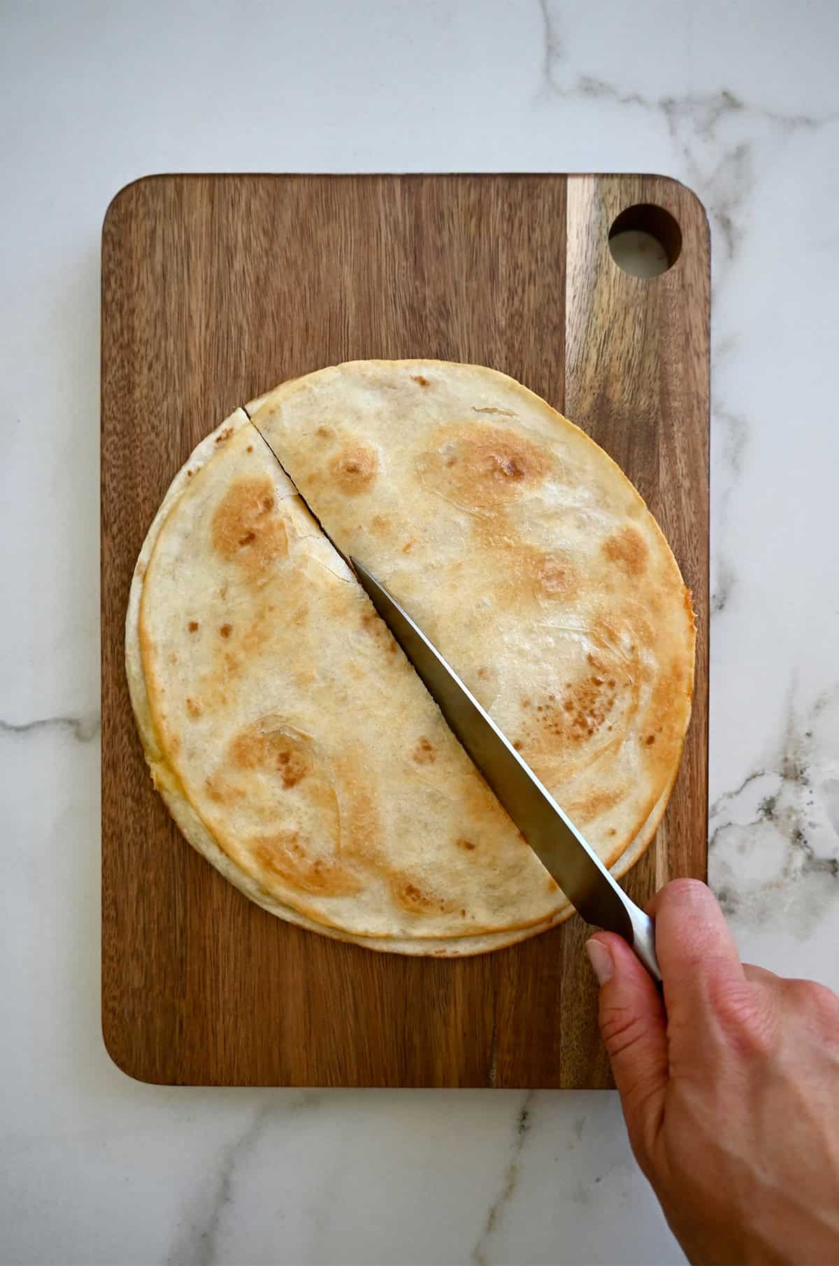 A hand holidng a sharp knife cuts a cheese quesadilla on a cutting board.