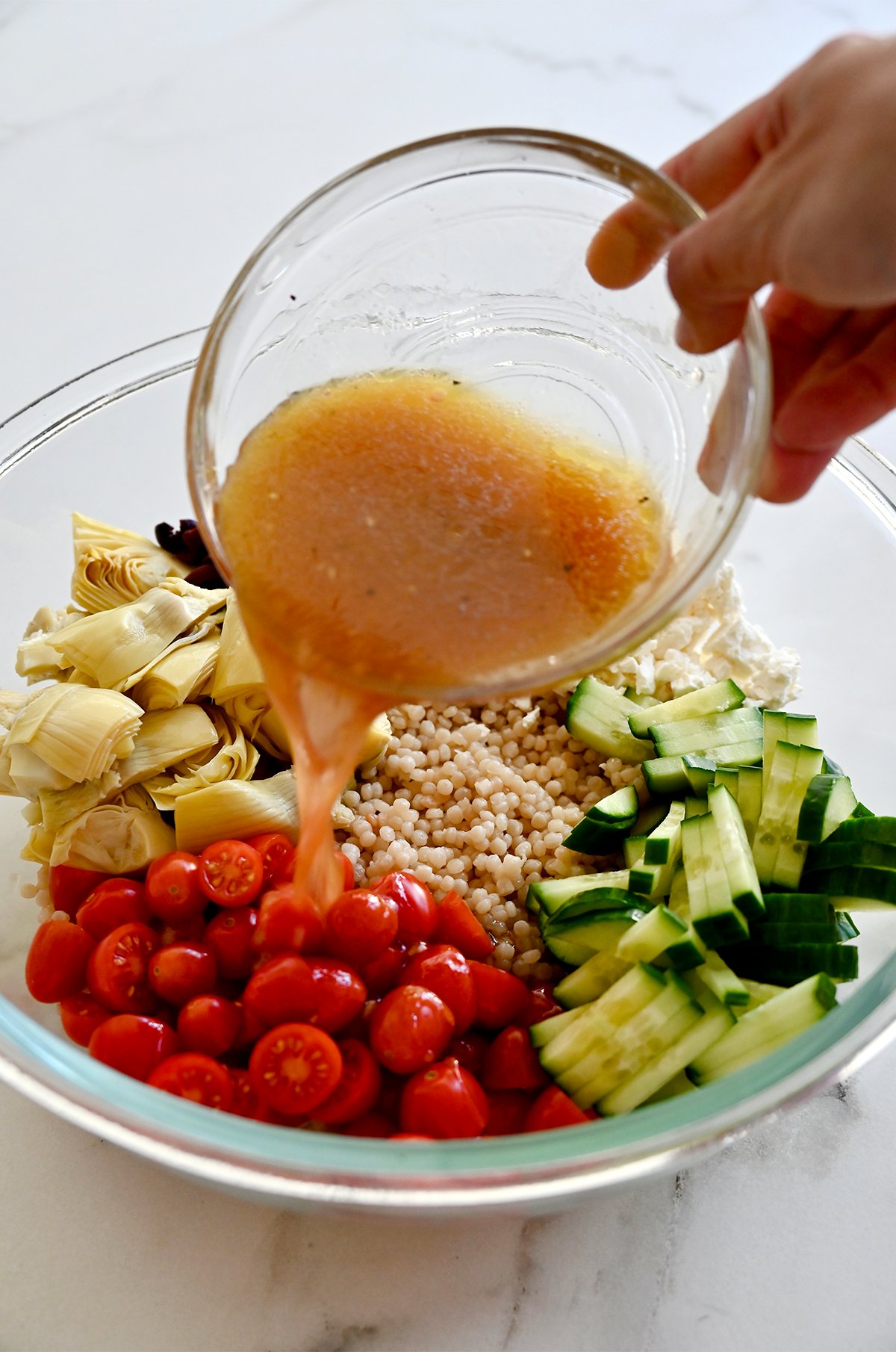 Pouring red wine dressing from a small atop couscous, halved cherry tomatoes, sliced cucumbers, chopped artichoke hearts, sliced kalamata olives and crumbled feta in a large bowl.