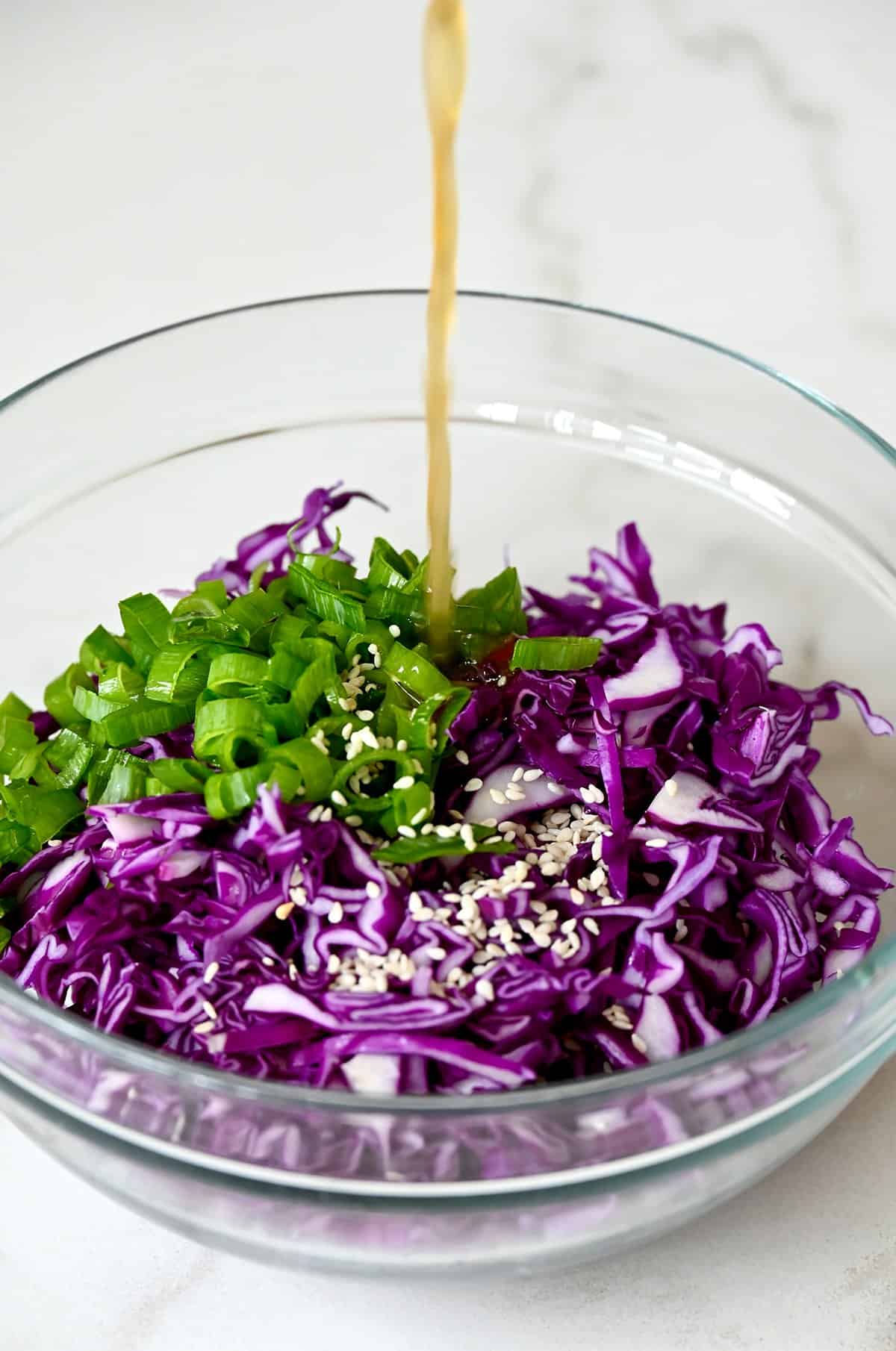 Asian dressing being poured over shredded purple cabbage, chopped scallions and sesame seeds in a large bowl.