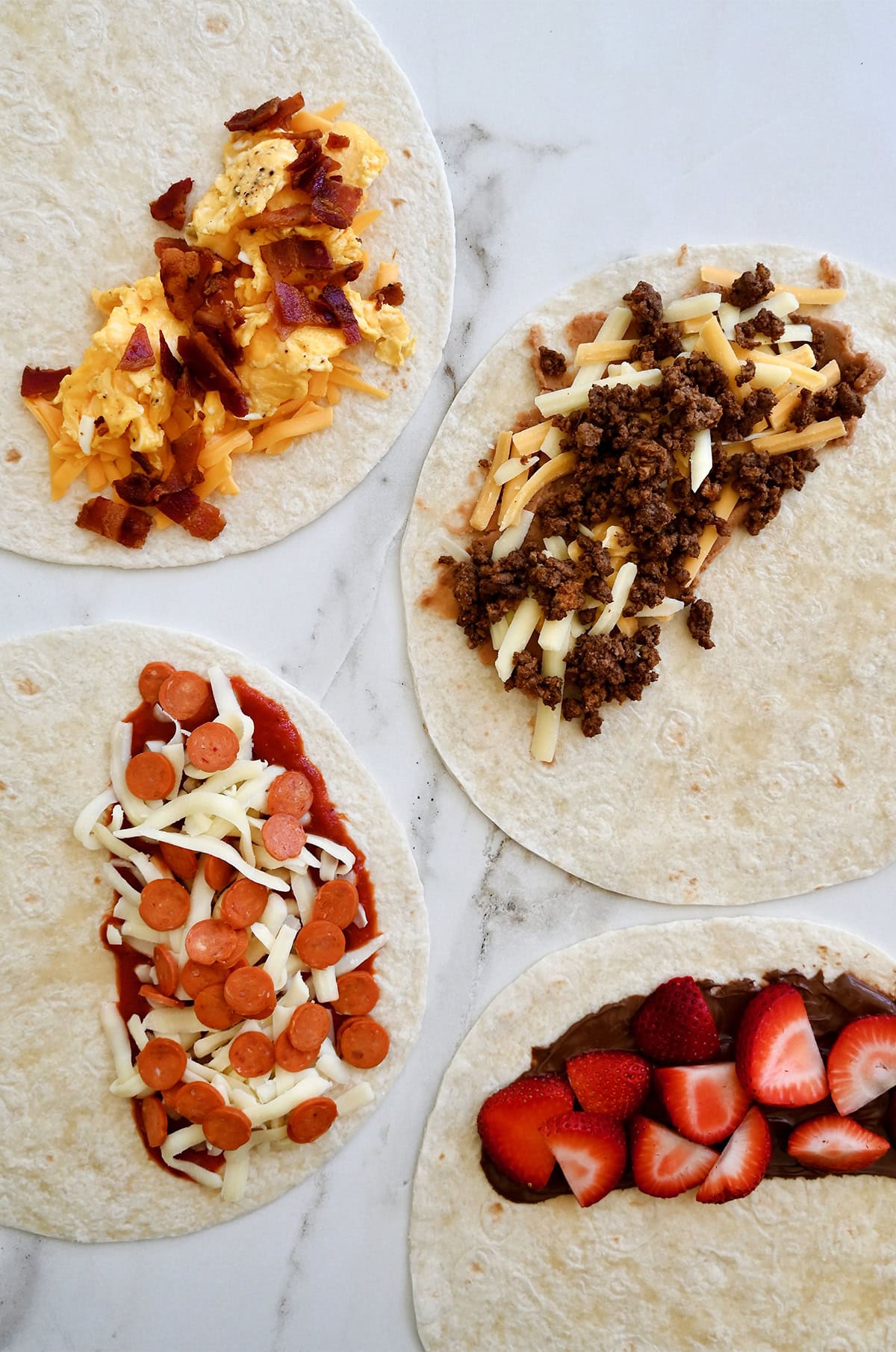 Four tortillas topped with different quesadilla fillings: scrambled eggs with bacon, taco-seasoned ground beef with cheese, Nutella with sliced strawberries, and marinara sauce with mini pepperoni.