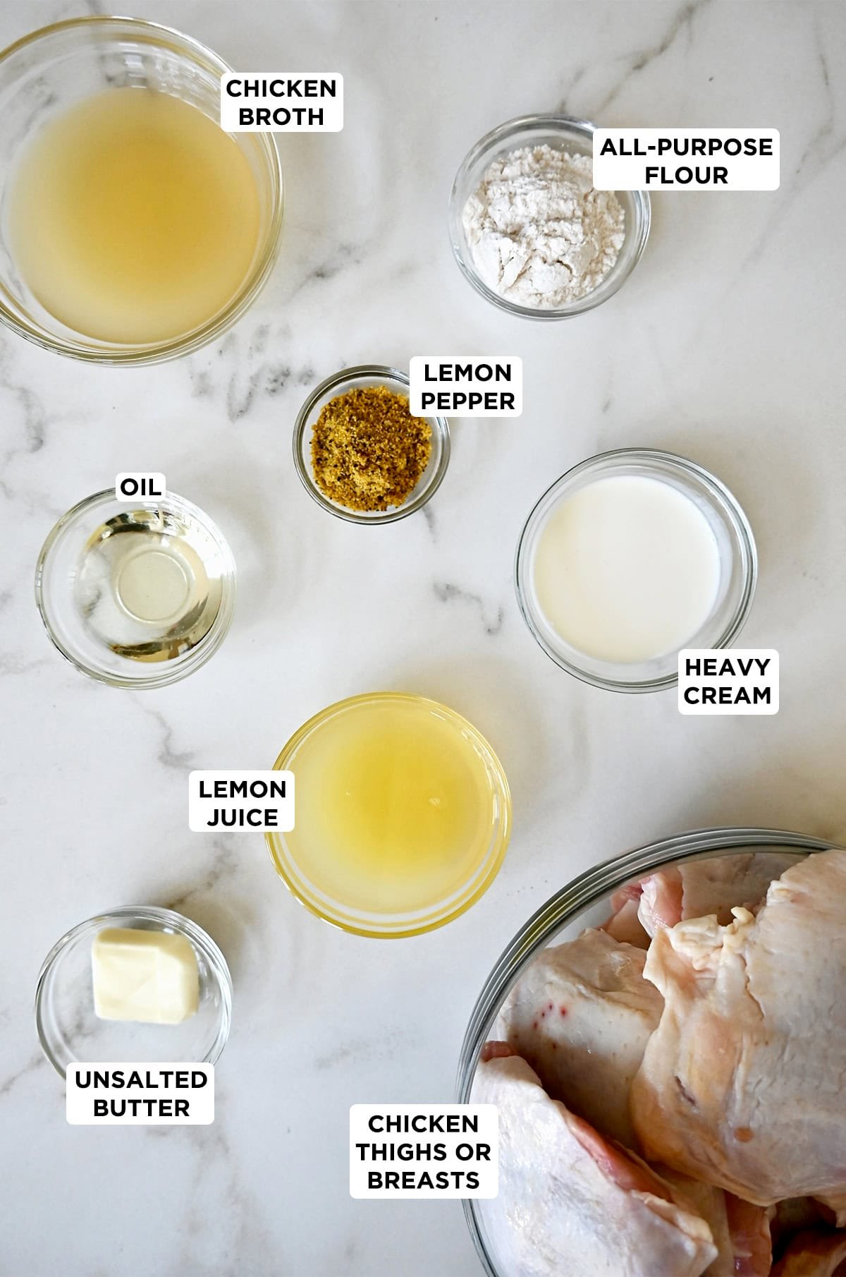Ingredients to make lemon pepper chicken in various sizes of glass bowls, including boneless, skin-on chicken thighs, lemon pepper seasoning, fresh lemon juice, butter, flour, chicken broth, and heavy cream.