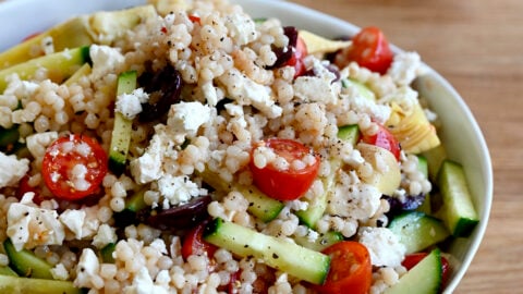 Mediterranean couscous salad with sliced cucumbers, cherry tomatoes, Kalamata olives, artichoke hearts and feta cheese, all tossed in a red wine vinegar and lemon juice vinaigrette in a large serving bowl.