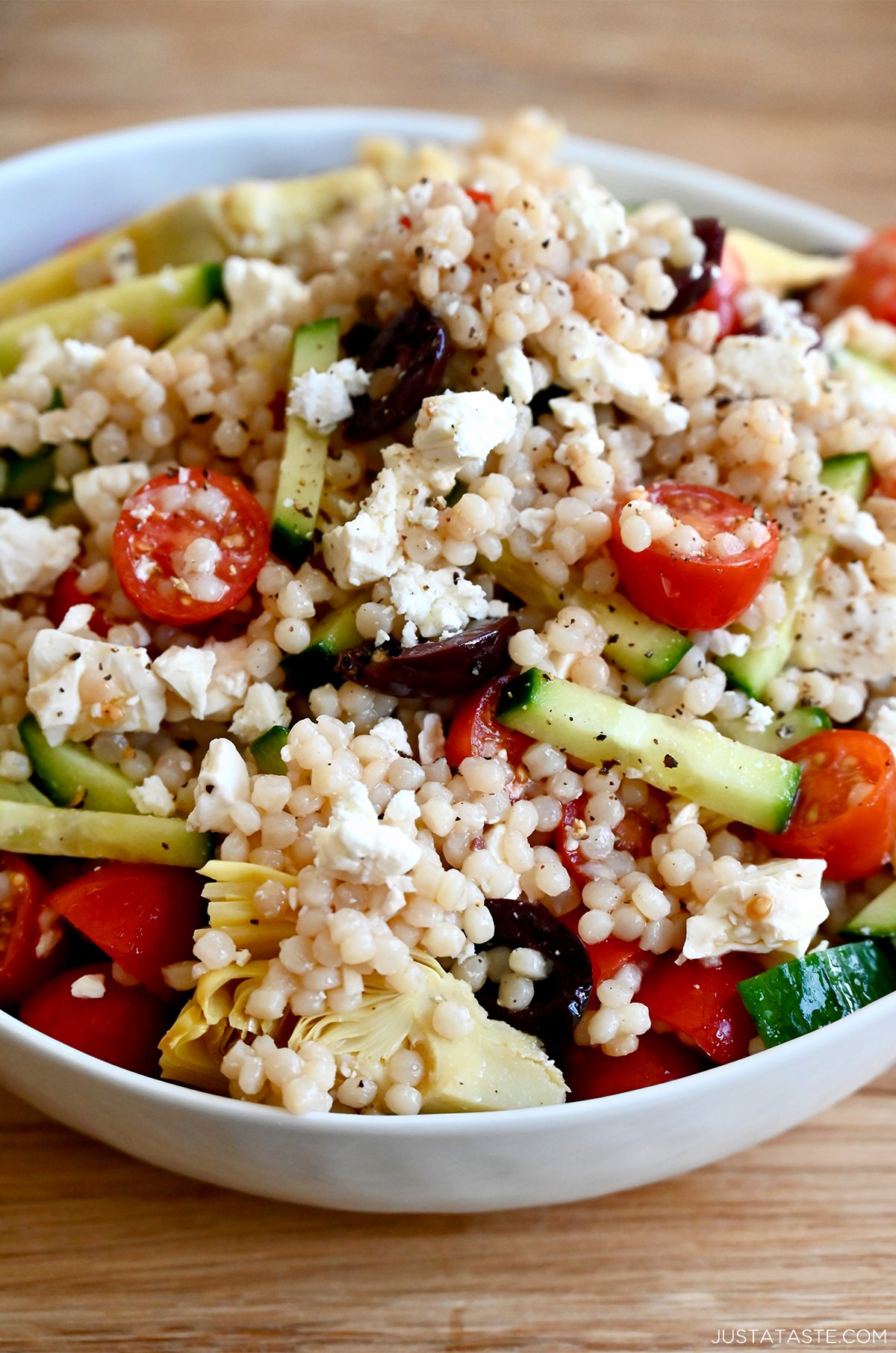 Mediterranean couscous salad with a red wine vinaigrette, cucumbers, tomatoes, kalamata olives and crumbled feta cheese in a large white serving bowl.