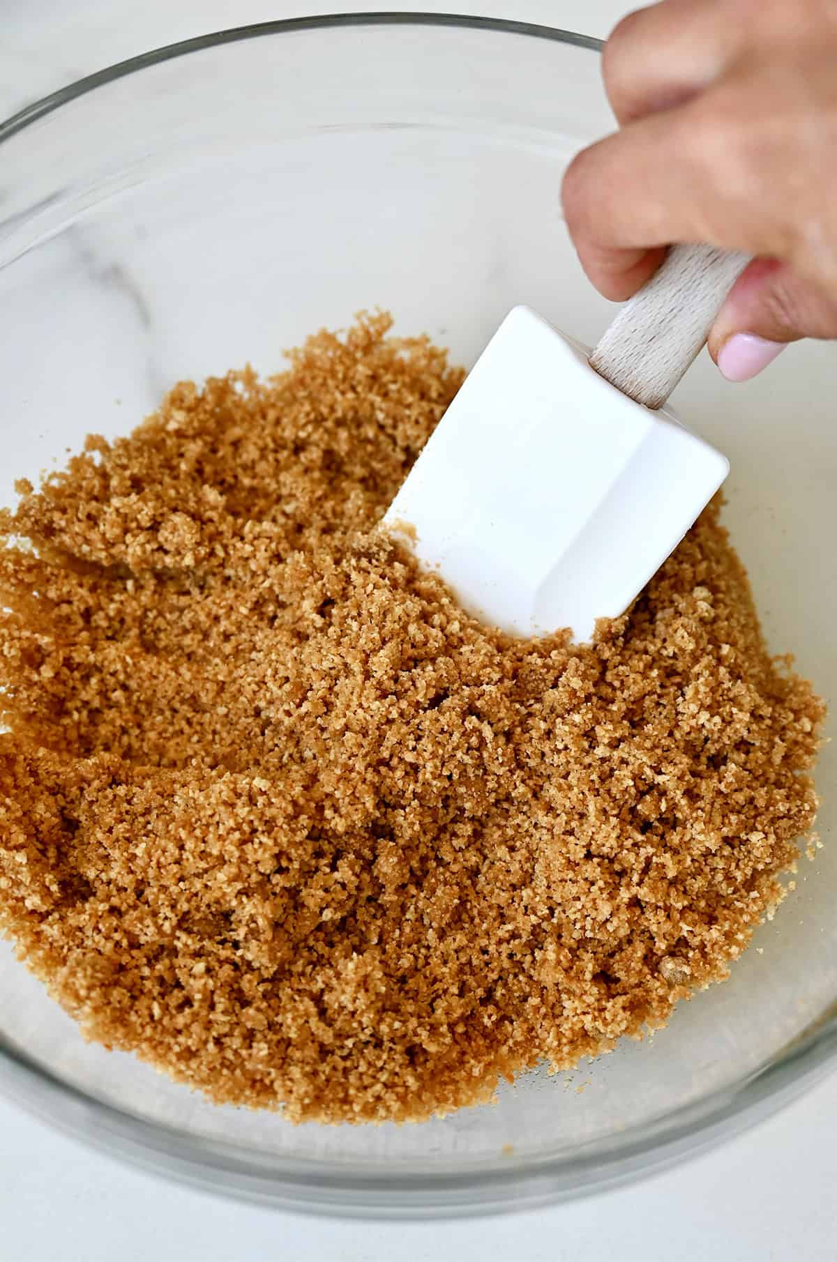 Graham cracker, melted butter and brown sugar mixture in a bowl with a spatula. The mixture is coarse and sandy.