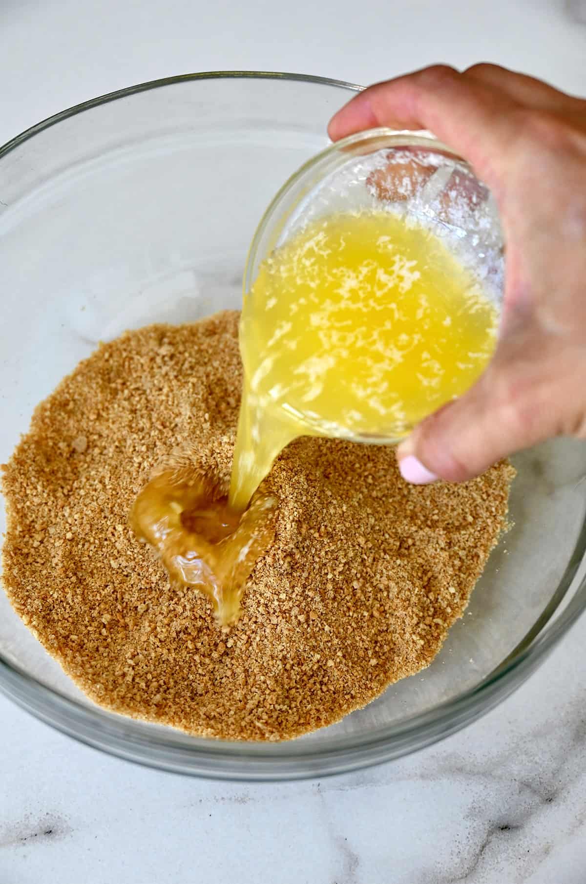 Melted butter being poured from a small bowl into a larger bowl with graham cracker crumbs.