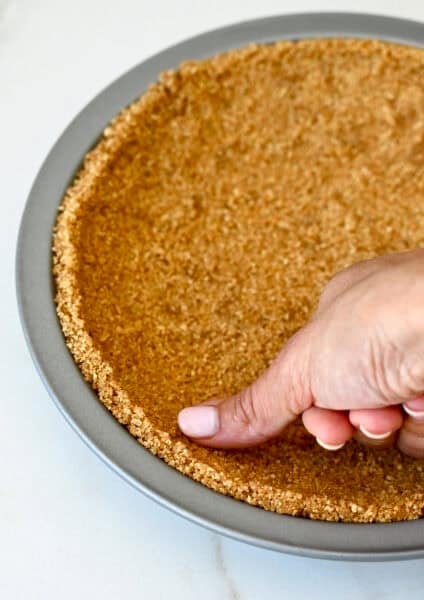 A hand presses a crushed graham cracker, melted butter and brown sugar mixture into a pie plate to make a graham cracker crust.
