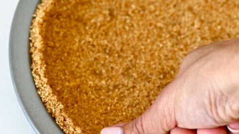 A hand presses a crushed graham cracker, melted butter and brown sugar mixture into a pie plate to make a graham cracker crust.